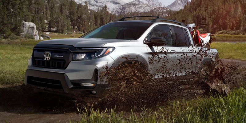 An all-new 2024 Gray Honda Ridgeline hauling a dirt bike in the bed, mudding through a puddle in the mountains. - Boyd Honda Oxford, NC