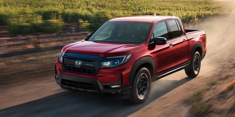 Driver's side view of a red 2025 Honda Ridgeline driving down a dirt road at dusk