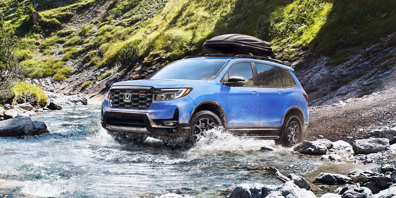 A blue Honda Passport SUV driving through a shallow river in a lush, rocky, and forested landscape.