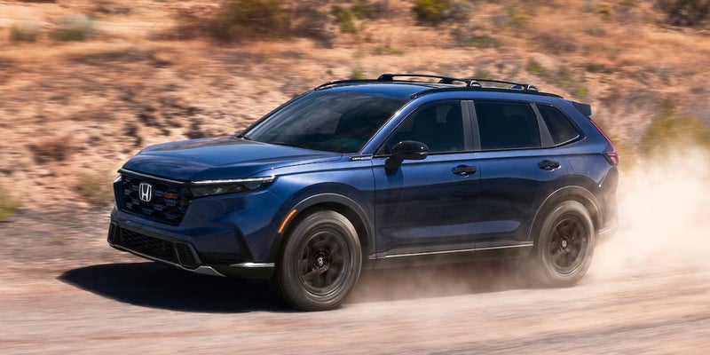 Blue Honda CR-V SUV driving on a dusty dirt road through a rugged desert landscape.